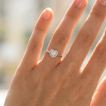 Close-up of a hand wearing a silver heart-shaped diamond ring on the ring finger, with a halo of small stones.