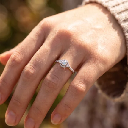 Close-up of a hand wearing a diamond halo engagement ring on a bare finger outdoors.