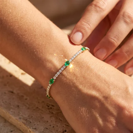 A delicate gold bracelet with emerald stones and embedded diamonds on a wrist, glinting in sunlight as a hand touches it