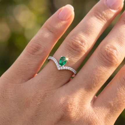 Close-up of a silver ring with a pear-cut emerald and diamond-studded band on a finger, sunlight sparkle on the gem.