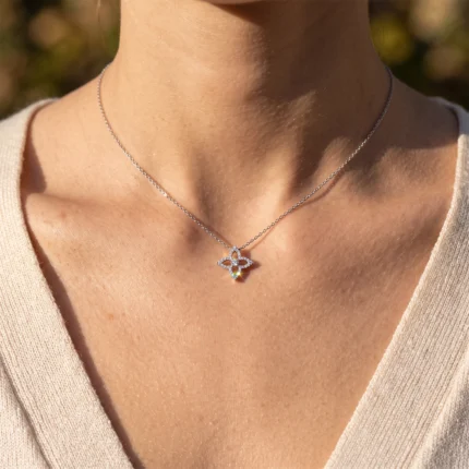 Close-up of a delicate silver chain necklace with a four-leaf-clover pendant and a rainbow teardrop gem resting on the collarbone of a person wearing a beige top.