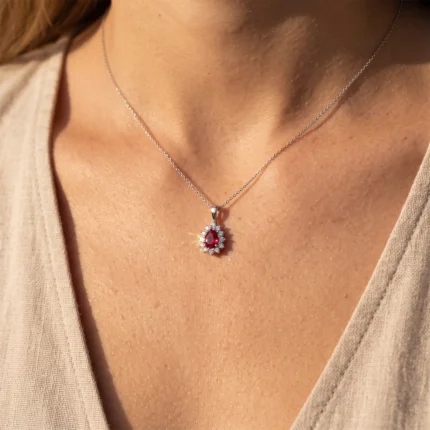 Silver necklace with a red teardrop gemstone pendant surrounded by small clear stones on a delicate chain.