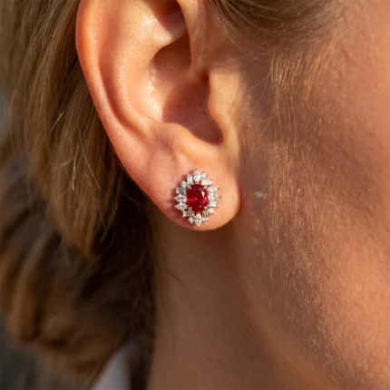 Close-up of an ear with a ruby-and-diamond halo earring featuring a central red ruby surrounded by clear diamonds in a sunlit setting.