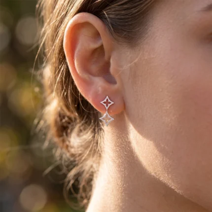 Close-up of a woman's ear wearing a two-piece diamond-shaped star jewelry earring set. The top small diamond star connects to a longer dangling diamond star pendant.