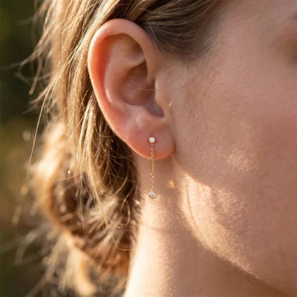 Close-up of a right ear wearing a gold drop earring with a small diamond stud and a delicate hanging chain.