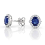 Oval blue sapphire stud earrings surrounded by a halo of diamonds set in white metal (likely white gold).