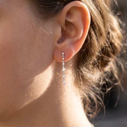 Close-up of a woman's ear wearing a long dangling diamond earring with multiple small stones in a vertical line, illuminated by sunlight.