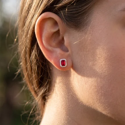 Close-up of a woman's ear with a rectangular red ruby stud surrounded by a diamond halo on the earlobe.