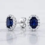 Pair of silver earrings with oval dark-blue sapphires surrounded by a halo of diamonds on a light marble surface.
