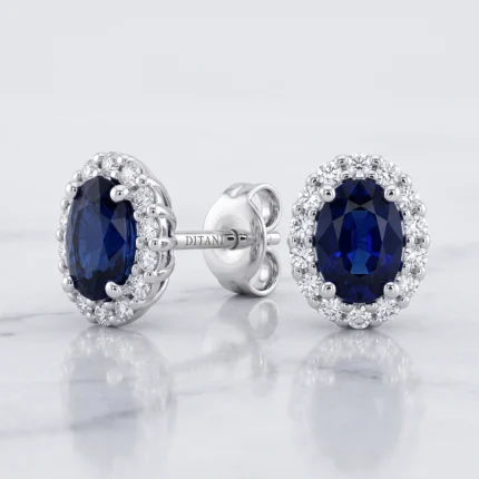 Pair of silver earrings with oval dark-blue sapphires surrounded by a halo of diamonds on a light marble surface.