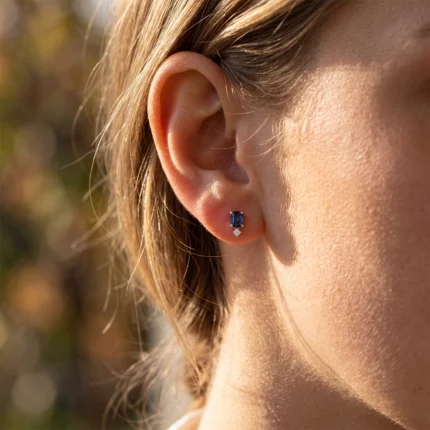 Close-up of a woman's ear with a blue rectangular gemstone stud earring and a small diamond below.