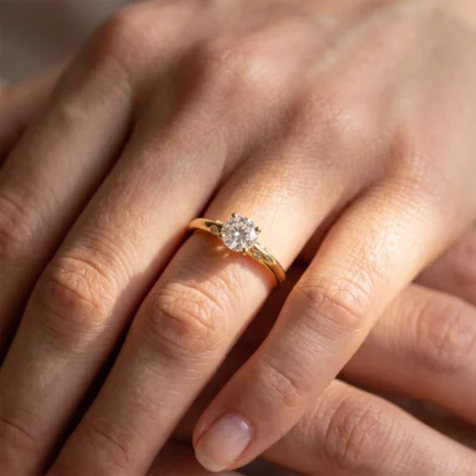 Close-up of a gold ring with a round-cut diamond on a finger, in a four-prong setting, showing a solitaire engagement ring.