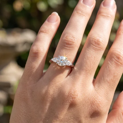 Close-up of a hand wearing a diamond engagement ring with a large center stone and two side stones on a gold band outdoors.