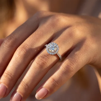 Close-up of a halo engagement ring on a finger, featuring a center round diamond surrounded by a halo of smaller stones and a pavé band.
