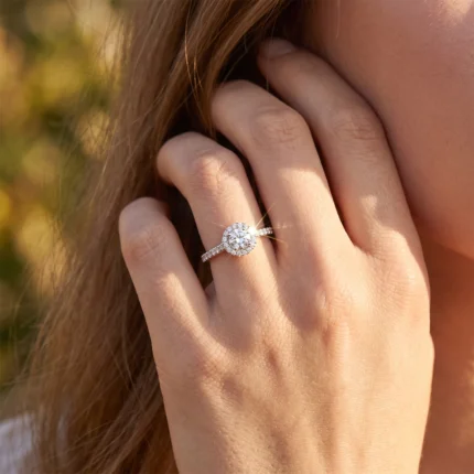 Close-up of a hand showing a diamond halo engagement ring on the ring finger, sunlight catching the stones.