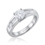 Close-up of a solitaire diamond engagement ring with a round center stone and baguette side stones in a white metal band in a prong setting on a white background.