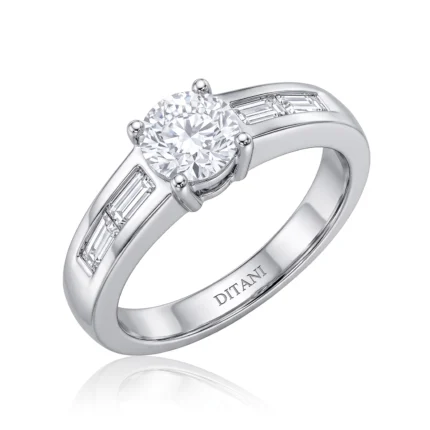Close-up of a solitaire diamond engagement ring with a round center stone and baguette side stones in a white metal band in a prong setting on a white background.