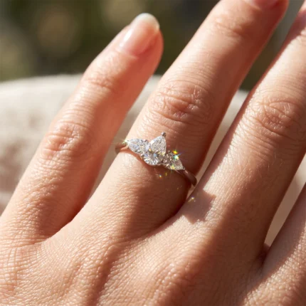 Close-up of a hand wearing a diamond engagement ring with a pear-cut center stone and two side marquise diamonds, sunlight glinting.