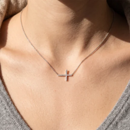 Silver necklace with a small, diamond-studded cross pendant resting on the collarbone area of a person wearing a gray top.