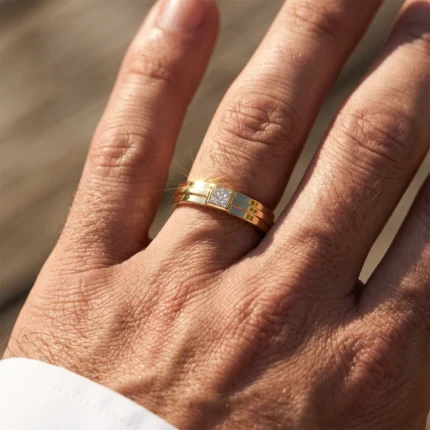 Close-up of a gold two-band ring with a square diamond centerpiece worn on a man's ring finger, sunlight glinting off the gem