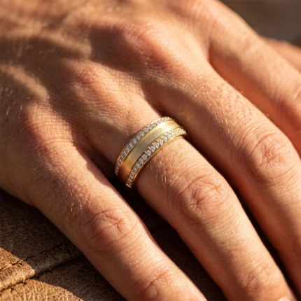 Close-up of a hand wearing a gold ring with two pavé-diamond bands on the ring finger, sunlight highlighting the stones and metal.