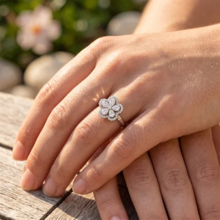 Close-up of a diamond flower ring on a woman's left ring finger outdoors, with a blurred floral background.