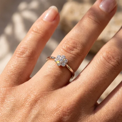 Close-up of a hand wearing a gold ring with a flower-shaped cluster of diamonds on the ring finger in sunlight.