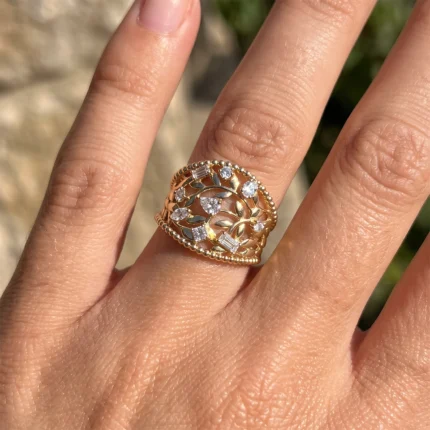 Rose-gold ring with an intricate leaf design and scattered clear stones, worn on a finger.