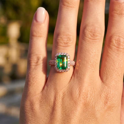 Close-up of a hand wearing an emerald-cut green gemstone ring with a diamond halo and pavé band on the finger.
