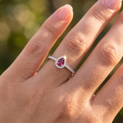 Close-up of a hand wearing a silver ring with a pear-shaped pink gemstone surrounded by diamonds on a thin pavé band, in sunlight.