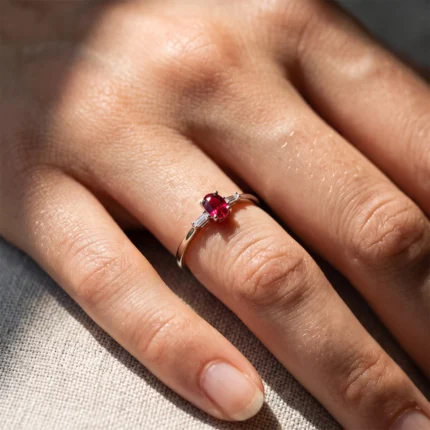 Close-up of a finger wearing a silver ring with a red ruby and small diamonds on each side.
