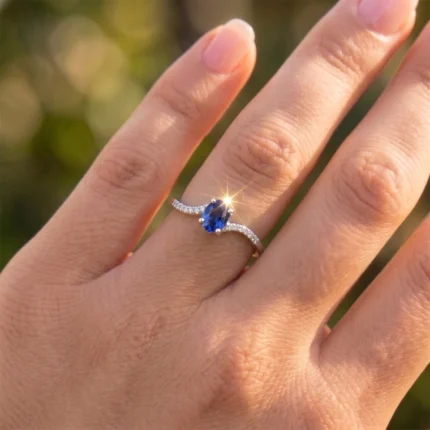 Close-up of a hand wearing a silver ring with an oval blue sapphire and diamond-studded band on the ring finger.