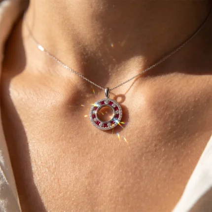 Close-up of a silver circular pendant with red gemstones on a delicate chain resting on the neck; sun glare adds sparkle.