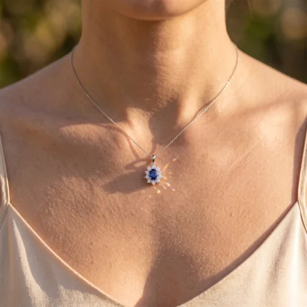 Close-up of a woman's neck wearing a silver chain necklace with a blue sapphire pendant surrounded by small diamonds, outdoors in warm light.