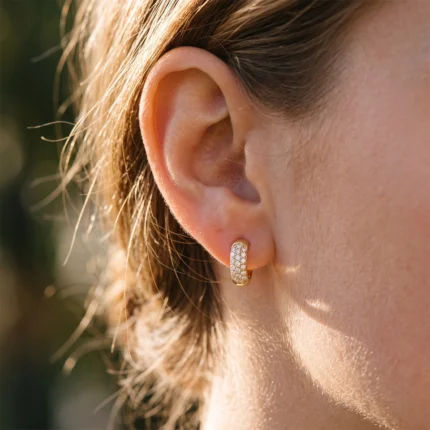 Close-up of a woman's left ear wearing a diamond-studded gold hoop earring outdoors.