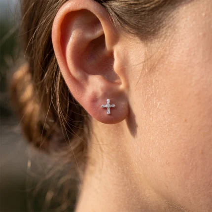 Close-up of a pierced earlobe adorned with a small silver cross-shaped stud earring jeweled with rectangular stones.