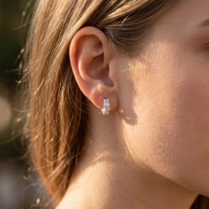Close-up of a woman's ear wearing a square, diamond-studded hoop earring catching sunlight.