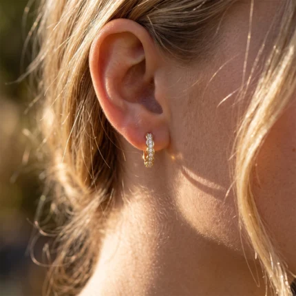 Close-up of a woman's ear with a small diamond-studded gold hoop earring on the earlobe, sunlight highlighting the jewelry.