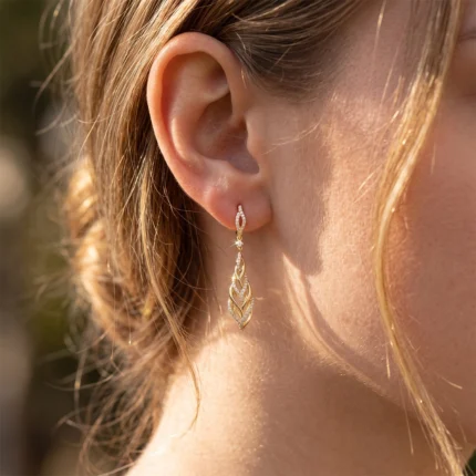 Gold leaf-shaped dangle earring with diamond accents on a earlobe, close-up view of jewelry detail on a woman's ear.