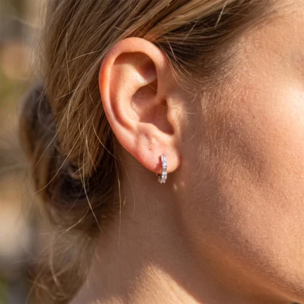 Close-up of a female ear wearing a small silver diamond hoop earring in sunlight.