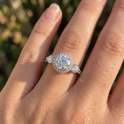 Engagement ring with a large central diamond in a halo setting and smaller side stones on a sunlit finger, close-up.