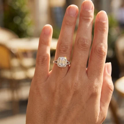 Hand wearing a diamond halo ring on the ring finger, outdoors; center emerald-cut stone with pavé halo diamonds.