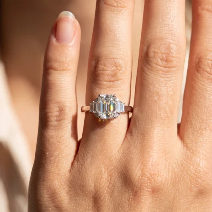 Close-up of a hand wearing an emerald-cut diamond engagement ring with a halo and baguette side stones on a warm-toned finger.