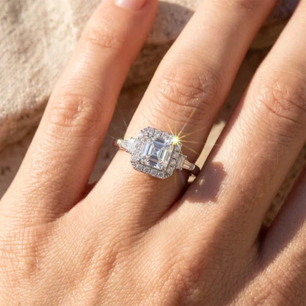 Close-up of a diamond engagement ring on a finger: an emerald-cut center stone with a halo of small diamonds and baguette side stones catching sunlight.