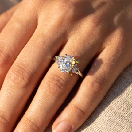 Engagement ring with a large oval diamond and side diamond accents on a finger, sunlight causing rainbow reflections.