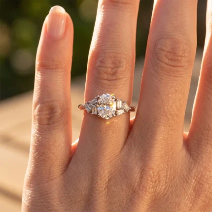 Engagement ring with a round center diamond and baguette side stones on a finger, illuminated by sunlight