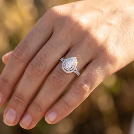 Close-up of a diamond engagement ring on the ring finger: pear-shaped center stone with double halo of small diamonds, sparkling in sunlight.