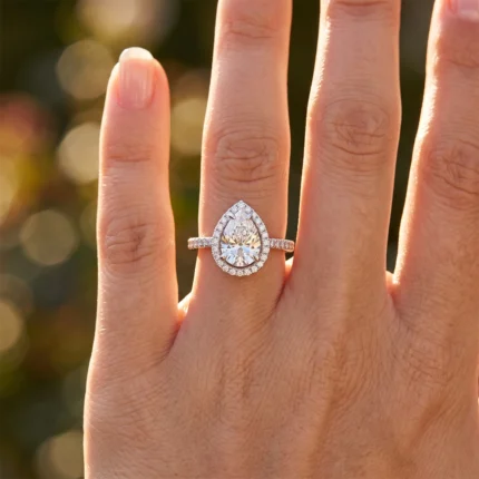Pear-cut diamond engagement ring with a diamond halo and pavé band on the ring finger, warm sunset backdrop.