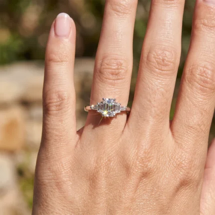 Close-up of a left hand wearing a silver ring with a large emerald-cut diamond and smaller side stones, sparkling in sunlight on the ring finger.