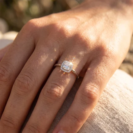 Close-up of a hand wearing a cushion-cut diamond engagement ring with a halo of smaller stones and a gold band, sparkling in sunlight.